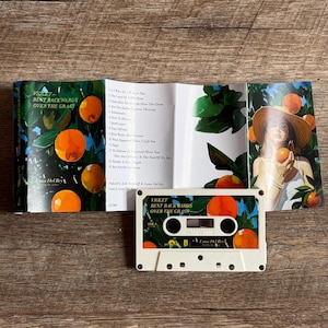 May include: A white cassette tape with the text "VIOLET BENT BACKWARDS OVER THE GRASS" and orange illustrations. The cassette case is open, revealing a booklet with song titles and artwork featuring oranges and a woman in a hat.