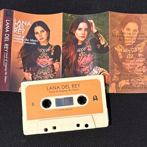 May include: A Lana Del Rey cassette tape titled Tired of Singing the Blues with a beige shell and orange label. The unfolded J-card features a portrait of the artist in a floral top, tracklist, and the text Lana Del Rey Tired of Singing the Blues.