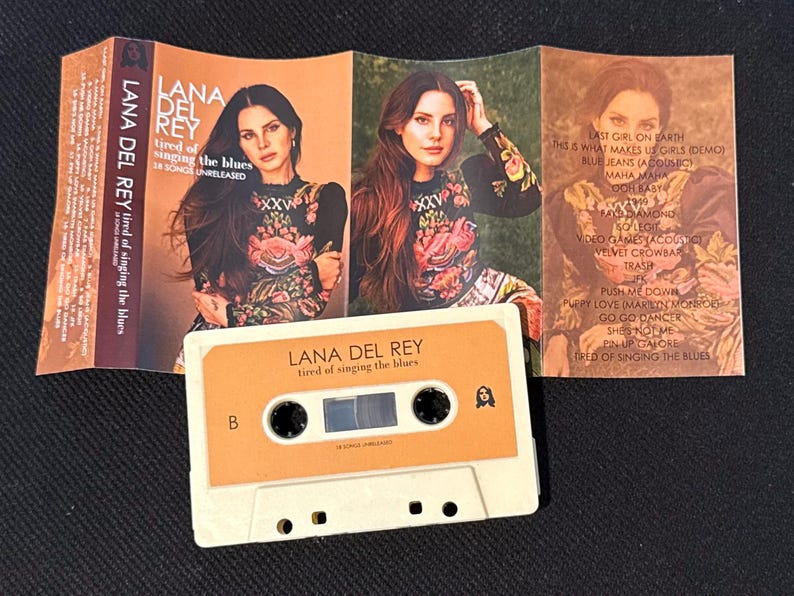 May include: A Lana Del Rey cassette tape titled Tired of Singing the Blues with a beige shell and orange label. The unfolded J-card features a portrait of the artist in a floral top, tracklist, and the text Lana Del Rey Tired of Singing the Blues.