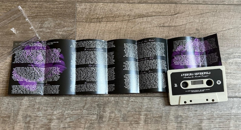 May include: A cassette tape and its insert are displayed. The cassette is off-white with black text and a black label. The insert is black with white text and purple floral accents. The cassette reads "Avenged Sevenfold" and "Waking the Fallen" on the label.