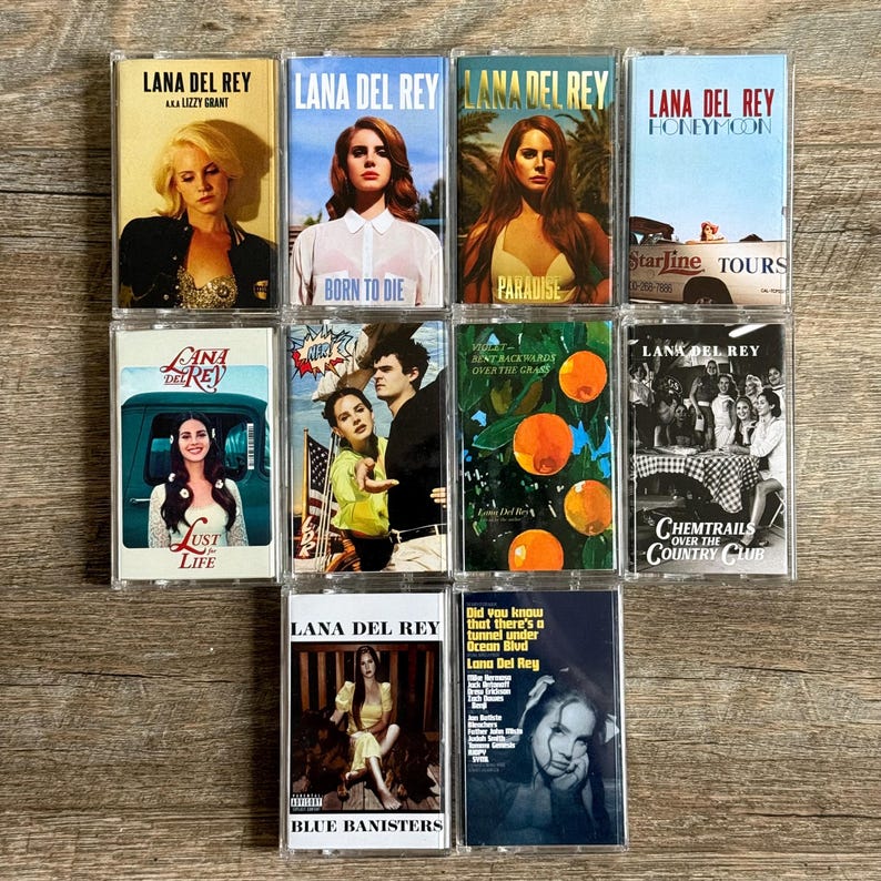 May include: A collection of ten Lana Del Rey cassette tapes. The album covers feature various images of the artist, with titles including "Born to Die," "Paradise," and "Blue Banisters." The cassettes are displayed on a wooden surface.