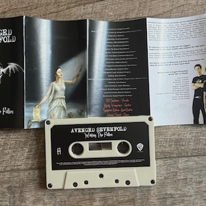 May include: A cassette tape and album booklet for the band Avenged Sevenfold's album, "Waking the Fallen." The cassette is off-white with black text. The booklet features band photos and lyrics.