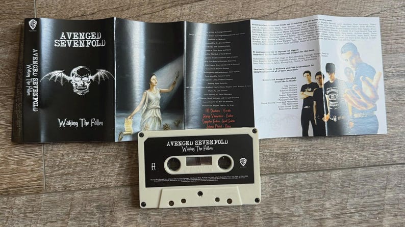 May include: A cassette tape and album booklet for the band Avenged Sevenfold's album, "Waking the Fallen." The cassette is off-white with black text. The booklet features band photos and lyrics.