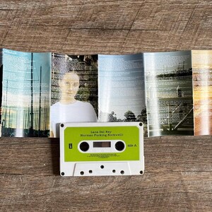 May include: A vintage cassette tape labeled "Lana Del Rey Norman Fucking Rockwell" with a green and white design. The cassette is placed in front of a folded album cover with images of people and landscapes.