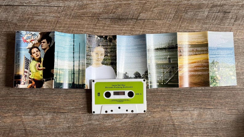 May include: A vintage cassette tape labeled "Lana Del Rey Norman Fucking Rockwell" with a green and white design. The cassette is placed in front of a folded album cover with images of people and landscapes.