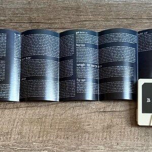 May include: A white cassette tape labeled "Avenged Sevenfold Nightmare" with a black and gray design. The cassette is next to a folded booklet with black pages and white text. The items are on a wooden surface.