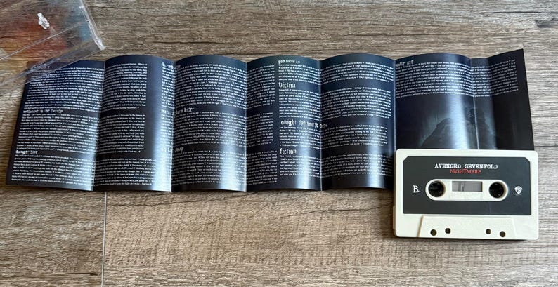 May include: A white cassette tape labeled "Avenged Sevenfold Nightmare" with a black and gray design. The cassette is next to a folded booklet with black pages and white text. The items are on a wooden surface.