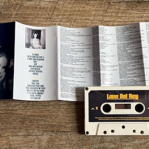 May include: A Lana Del Rey cassette tape with the album title "Did you know that there's a tunnel under Ocean Blvd" and lyrics. The cassette is white with a black and gold label. The album cover is visible.