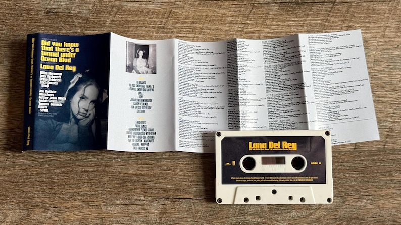 May include: A Lana Del Rey cassette tape with the album title "Did you know that there's a tunnel under Ocean Blvd" and lyrics. The cassette is white with a black and gold label. The album cover is visible.
