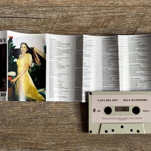 May include: A cassette tape and album booklet for Lana Del Rey's "Blue Banisters." The booklet features photos of the artist in a yellow dress. The cassette is light pink with the album title printed on it.