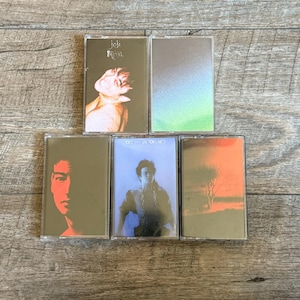 May include: Five cassette tapes featuring album art. The top left tape has the name "joji" and a figure. The other tapes have different images, including a person's face, a person in a blue outfit, and an abstract landscape. The tapes are in clear plastic cases.