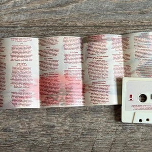 May include: A vintage cassette tape with a white case and a folded booklet. The tape has the words "Lana Del Rey" printed on it in red. The booklet is filled with small red text. The cassette tape and booklet are on a wooden surface.