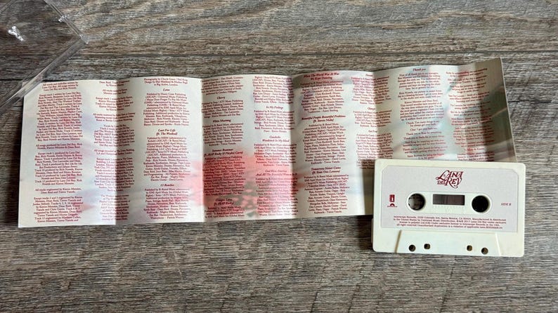 May include: A vintage cassette tape with a white case and a folded booklet. The tape has the words "Lana Del Rey" printed on it in red. The booklet is filled with small red text. The cassette tape and booklet are on a wooden surface.