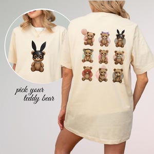 May include: A cream-colored t-shirt featuring a graphic of a teddy bear wearing black bunny ears on the front and a grid of nine different teddy bears on the back. The text pick your teddy bear is printed in black script on the lower left side.
