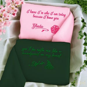 May include: Two folded fabric items, one pink and one dark green, are nestled in a basket. The pink item has the text "I know I'm who I am today because I knew you" and "Glinda" embroidered in pink. The green item has the text "you'll be with me like a handprint on my heart" and "Elphaba" embroidered in green.