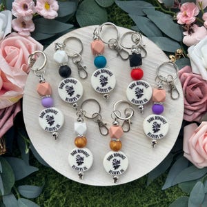 May include: A selection of keyrings with white, circular charms displaying the text "DARK ROMANCE READERS UK" and a floral motif. Each keyring features colourful beads and a silver-tone clasp.