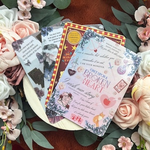 May include: A collection of decorative bookmarks with various designs and text. The bookmarks feature illustrations of a woman, butterflies, roses, and text such as "Once Upon a Broken Heart" and "I am a monster". The color palette includes pink, red, and blue.