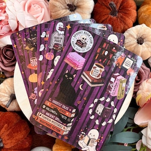 May include: A rectangular sticker sheet featuring a purple and black striped background with illustrations of a black cat, ghosts, stacks of books, a mug labelled Books, Brews, & Cozy Views, and a circular Spooky Book Club Madrid Spain Est. 2023 logo.