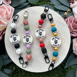 May include: Four beaded keychains with black lobster clasps. Each keychain features a white circular charm with the text "DARK ROMANCE READERS UK" and a floral design. The beads are in various colours, including black, white, pink, green, and gold.