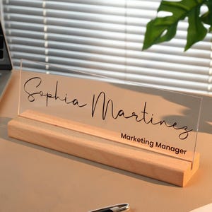 May include: A clear acrylic desk nameplate with the name "Sophia Martinez" and the title "Marketing Manager" in black script. The nameplate sits on a light wood base. A black pen and a laptop are also on the desk.