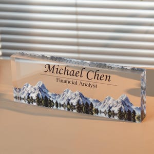 May include: A clear acrylic desk nameplate with the name "Michael Chen" and the title "Financial Analyst" printed in black. The nameplate features a mountain range design at the bottom. The nameplate is rectangular and sits on a desk.
