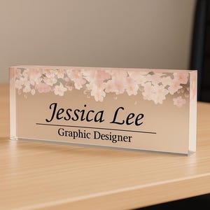 Könnte beinhalten: Rechteckiges Namensschild mit floralem Design und dem Namen "Jessica Lee" sowie dem Titel "Grafikdesigner" in schwarzer Schrift. Die Oberseite des Namensschildes zeigt einen Rand aus rosa und weißen Blüten.