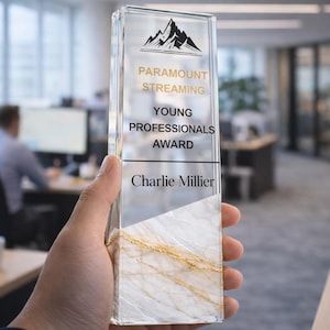 Puede incluir: Un premio rectangular de vidrio con una base de mármol y detalles dorados. El premio presenta el texto "Paramount Streaming Young Professionals Award" y el nombre del destinatario, Charlie Millier, debajo de un gráfico de montaña.