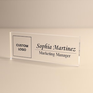Peut inclure: Plaque de bureau en acrylique transparent avec texte noir. Le c&ocirc;t&eacute; gauche comporte une case avec "CUSTOM LOGO" &agrave; l'int&eacute;rieur. Le c&ocirc;t&eacute; droit indique "Sophia Martinez" et "Marketing Manager" en dessous. La plaque est rectangulaire.