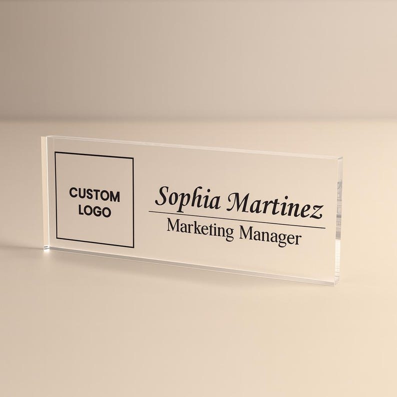 Peut inclure: Plaque de bureau en acrylique transparent avec texte noir. Le c&ocirc;t&eacute; gauche comporte une case avec "CUSTOM LOGO" &agrave; l'int&eacute;rieur. Le c&ocirc;t&eacute; droit indique "Sophia Martinez" et "Marketing Manager" en dessous. La plaque est rectangulaire.