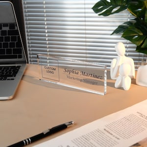 Peut inclure: Plaque de bureau en acrylique transparent avec le texte "Sophia Martinez, Marketing Manager". La plaque comprend &eacute;galement un espace pour un logo personnalis&eacute;. Un stylo noir et un ordinateur portable sont visibles en arri&egrave;re-plan.