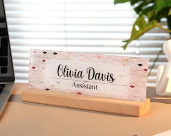 May include: A desk nameplate with the name "Olivia Davis" and the title "Assistant" printed in black on a clear acrylic panel. The panel has a decorative pattern and sits in a light wood base.