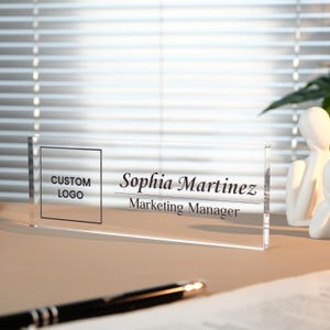 Peut inclure: Plaque de bureau en acrylique transparent avec le texte "Sophia Martinez, Marketing Manager". La plaque comporte &eacute;galement un espace pour un logo personnalis&eacute;. Un stylo noir et une figurine blanche sont en arri&egrave;re-plan.