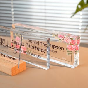 Peut inclure: Plaques nominatives en acrylique transparent avec des motifs de roses. Les plaques affichent des noms et des titres en texte noir. Une plaque est fix&eacute;e sur une base en bois. Le fond est de couleur neutre.