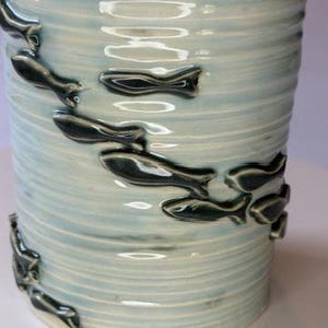 May include: A cylindrical ceramic vessel with a light blue, ribbed glaze finish. The exterior is decorated with a series of small, dark, raised fish motifs that appear to be swimming in a diagonal pattern across the textured surface of the container.