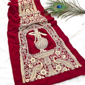 May include: A red velvet textile with intricate gold embroidery. The design features a central arch with a dancer figure, surrounded by floral and peacock motifs. The textile is likely a decorative piece.