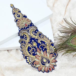 May include: A detailed, blue embroidered applique with gold, red, and yellow accents. The ornate design features floral and paisley patterns, embellished with rhinestones. The applique is likely for embellishing clothing or accessories.