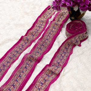 May include: Magenta fabric trim with detailed gold and blue embroidery and small, sparkling embellishments. One section is rolled up, others are laid out, displaying the intricate design. Suitable for craft projects. The trim is measured in centimetres.