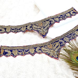Puede incluir: Dos piezas de tela bordada ornamentada con base azul marino y diseños florales dorados. Los bordes son festoneados y están ribeteados con tela roja. Una pluma de pavo real es visible en el fondo.