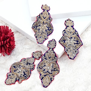 Può includere: Quattro toppe ricamate ornate con un design floreale e a foglie. Le toppe presentano filo dorato su sfondo blu navy, con bordi rossi e piccole paillettes. Le toppe sono disposte su una superficie bianca.