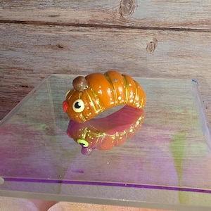 May include: A whimsical, orange caterpillar ring with a brown head and white and gold accents. The ring has a red mouth and a large, round eye. The ring is reflected in the surface below.