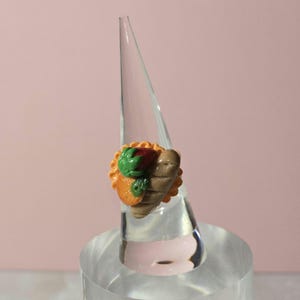 May include: A miniature taco ring with a brown, orange, and green design. The ring features a small taco with a brown tortilla, orange filling, green lettuce, and a red tomato. The ring is displayed on a clear acrylic stand.