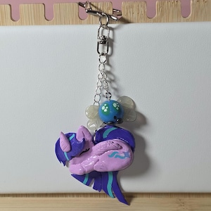 May include: A handcrafted keyring with a sleeping pony figurine in shades of purple and blue. The pony is suspended from a silver chain, with a blue and white bead, and a silver clasp. A unique accessory.
