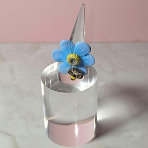Op de afbeelding: Een blauwe bloemenring met een gele kern en een kleine bijenaccent. De ring wordt gepresenteerd op een heldere acryl ringhouder met een roze basis. De achtergrond is zachtroze.