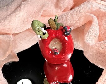 Puede incluir: Una caprichosa escultura de manzana roja con un centro ahuecado, adornada con una oruga verde, mariquitas y una pequeña criatura verde. La escultura se refleja en una superficie negra, con una tela color melocotón en el fondo.