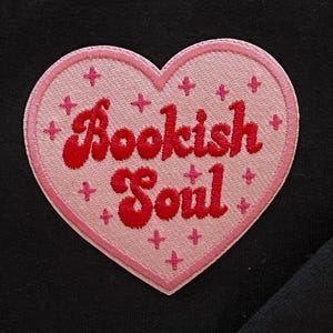 Op de afbeelding: Een roze hartvormige patch met de woorden "Bookish Soul" in het rood geborduurd. De patch heeft een roze rand en kleine roze kruisvormige accenten. De patch staat op een zwarte achtergrond.