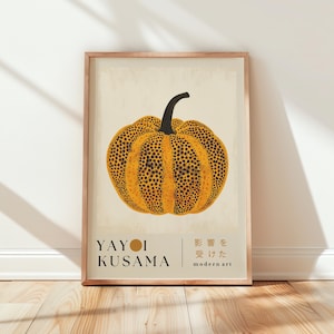 Puede incluir: Impresión artística enmarcada que representa una calabaza estilizada con un tallo negro y un cuerpo amarillo cubierto de puntos negros. La impresión incluye el nombre "YAYOI KUSAMA" y texto japonés, enmarcada en un marco de madera marrón claro.