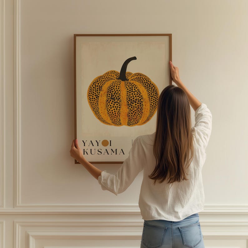 May include: A framed art print featuring a stylized pumpkin with a yellow and black spotted pattern. The artwork includes the text "YAYOI KUSAMA" at the bottom. The print is displayed on a white wall.