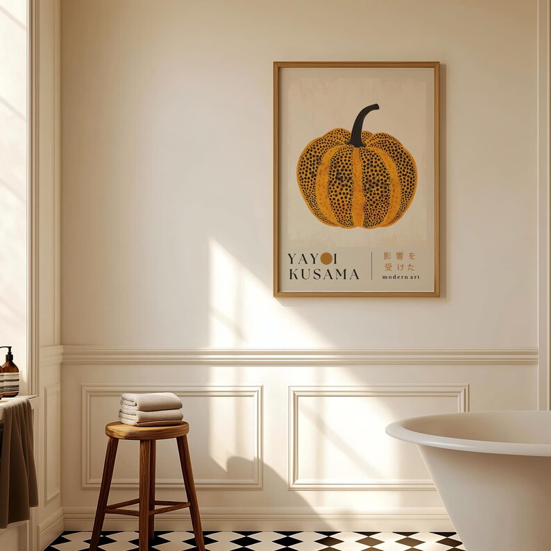 May include: A framed art print featuring a stylized orange pumpkin with black spots and a black stem. The print includes the text "YAYOI KUSAMA" and Japanese text. The artwork is displayed in a bathroom setting with a wooden stool and a bathtub.