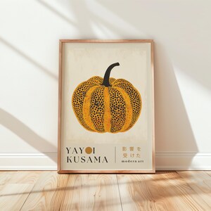May include: A framed art print featuring a stylized pumpkin with a black stem and a yellow body covered in black dots. The print includes the name "YAYOI KUSAMA" and Japanese text, framed in a light brown wooden frame.