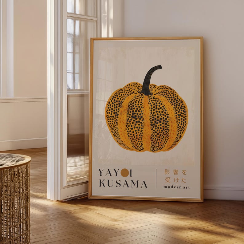 May include: A framed art print featuring a large, yellow pumpkin with black spots and a curved black stem. The print includes the artist's name, Yayoi Kusama, and Japanese text. The background is a light beige color.